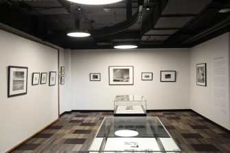 A gallery with several framed, black-and-white photographs along three walls. At the centre of the room is two glass display cases side-by-side, containing additional photos. 