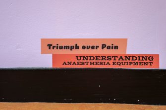 A purple wall with an illustration of two stacked book spines resting on a black skirting board. The titles read, "Triumph over Pain" and "Understanding Anaesthesia Equipment".