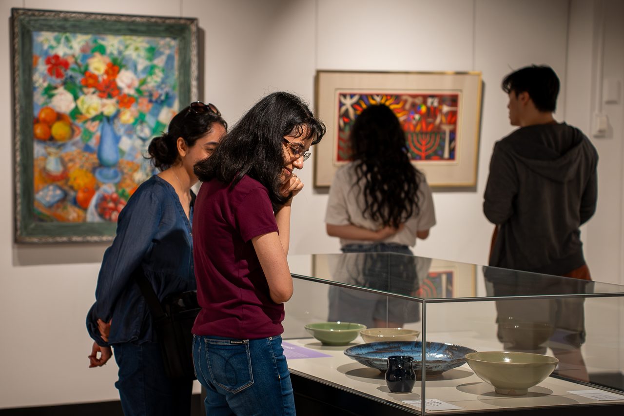 Four people view artworks in a gallery. On the left of the image, two people view ceramics in a display case. To the far right of the image, two people view paintings on the wall. 