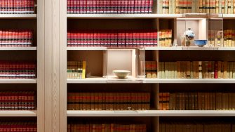 Display shelving wall containing mostly red or pale yellow coloured bound books and clear display boxes contatining sculputural artefacts.