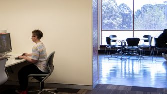 Side view of a person who is sitting in front of computer, white wall beside them. Distant communal tables seen through the open entrance way to the right. Trees seen through the full length windows with the outside daylight reflecting on the floor..