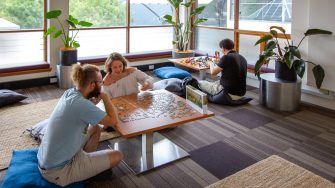  A room with large windows and indoor plants. 3 people sitting on cushions and low tables, doing recreational activities such as jigsaw puzzle and jenga blocks.