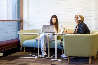  Students studying on a longer corner couch next to a window. Laptop tables in front of them as they use their laptops. One student with headphones on.
