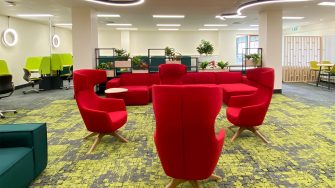 A study space in shades of green and white. Various types of group and individual study furniture are available, including sofas, lounge chairs, height adjustable desks and bar tables. Red sofas and high back lounge chairs in the centre. Line of open block shelving at different heights with potted plants behind and window in distance to one side. Curved narrow strip lighting on ceiling.