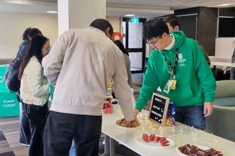 A Library Peer Mentor who is wearing a green hoodie uniform is talking to an event participant. There are cookies and chocolates spread on a table between them.