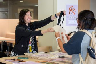A smiling Library staff member is handing out a Cover Crush mural merchandise to a student.