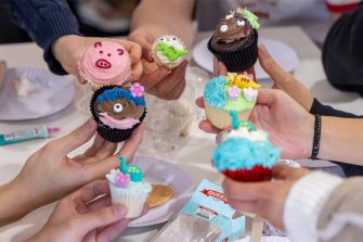 A close up of students' hands holding their cupcakes decorated with icing. The cupcakes are colourful, including a pink pig cupcake, chocolate monster cupcakes, and green and blue dinosaure cupcakes.