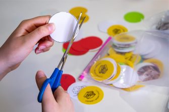 Pair of hands shown using blue handled scissors to cut circular white shape. Yellow, green and red circles, some with black book stack icon and text lie on the white table. A pile inside are inside a plastic bag next to a pink pencil.