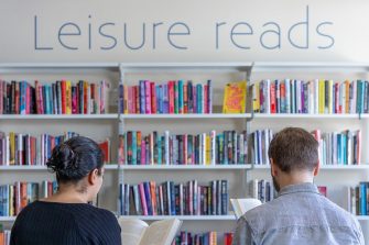 Two people seated next to each other are reading books. They are facing the UNSW Main Library Leisure reads display shelves.