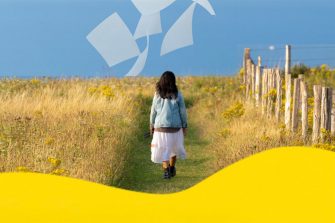 The back of a woman walking in a golden outback field. There are shapes that look like poetry pages in the blue sky.