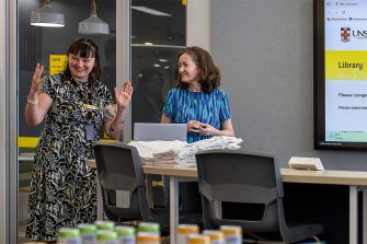 Two librarians smiling and presenting at a workshop. A digital screen next to them displays a Library page. Rows of canned drinks and stacks of UNSW Library merchandise are nearby.