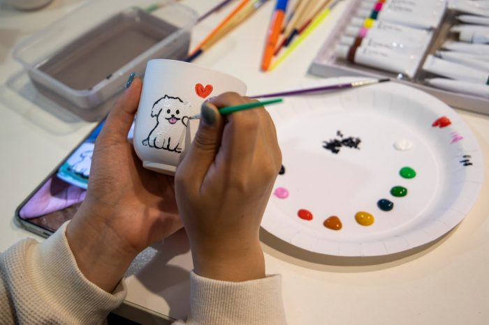  Close up of a student painting a cute dog image onto a mug. Paints and brushes are scatted on the table.