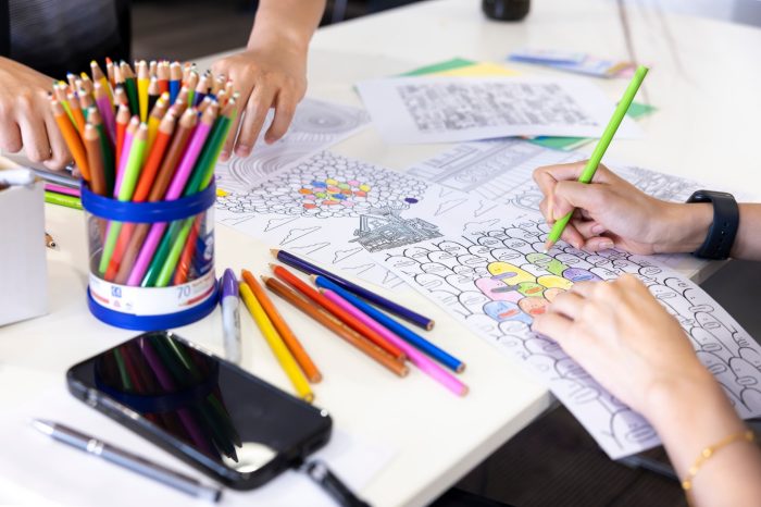  Close up of two students colouring in sheets of paper. There are coloured pencils and other colouring in sheets scattered on the table.