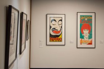 Photo of a gallery wall with two framed screen-printed artworks. The artwork to the left is a comic-style illustration of  a face with the word "NIMROD" below. The artwork to the right is a comic-style illustration of a woman's face with red hair and star earrings with the text "Sideshow in Burlesco, Nimrod".