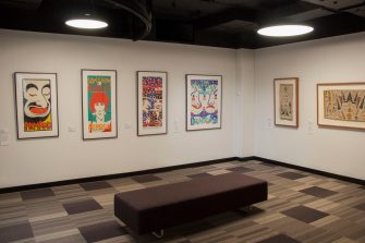A long shot photo of a gallery space, with two walls at right-angles. The left wall displays four framed large colourful illustrated artworks. The right wall displays two Australian Indigenous artworks with earthy colours. 