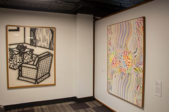 Photo of a gallery featuring two large artworks hanging on walls at right-angles. The artwork to the left is a bold, stylistic black and white illustration of a dining room scene. The artwork to the right shows detailed, abstracted patterns of various colours.