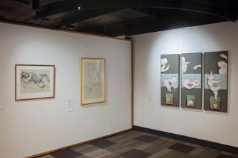 A photo of a gallery space, showing two adjacent walls with hanging artworks. The left wall has two wooden framed artworks with indiscernible black and white sketches. The right wall has a triptych of green coloured paintings with figures in various forms. 