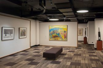 A long shot photo of a gallery space with several artworks hanging on the walls. To the right of the photo are two wooden sculptures of tall, crane-like birds.