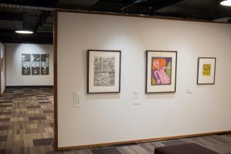 A long-shot photo of a gallery space with three framed artworks of different sizes mounted on the wall.  