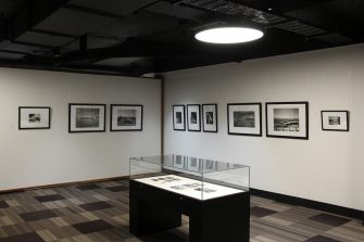 A corner of the exhibition gallery with nine black-and-white photographs along the two walls, and a glass display containing additional photos in the centre of the room.