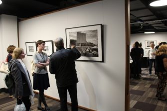 On opening night, several guests viewing and discussing a photo on display. One guest is gesturing towards a framed, black-and-white photograph. 