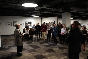 A women with silver hair and wearing a long coat. She is standing in front of the exhibition installation title wall, making a speech. Guests are looking towards her.