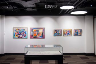 A gallery with five colourful, abstract paintings hanging on the wall. A glass display case is situated in the centre of the gallery space, in front of the paintings. 