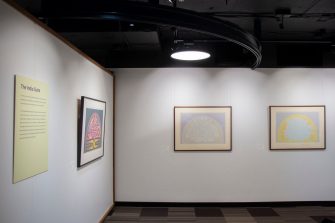 A gallery space with a view of two adjascent walls. Both walls display framed paintings with muted colours and soft shapes. 