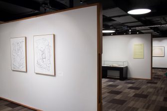 View of a gallery space across three separate walls. The wall closest to the camera shows two framed abstract drawings with a white background. 