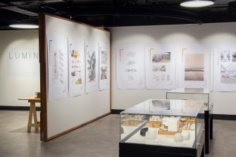 A photo of a gallery space, showing two adjacent walls with large posters hanging on them. To the lower right-hand of the image is a display case housing architectural models.
