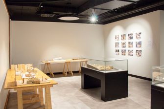 A photo of a gallery space with two wooden tables against the walls. Upon the tables are three-dimensional architectural models. To the right of the image is a wall with twelve colourful, but indiscernible, images mounted in a neat grid pattern. 