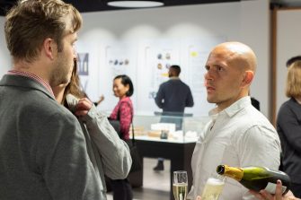 Photo with a man in a white shirt facing towards the camera, in conversation with another man with his back to the camera. In the background other guests mingle amonst the gallery display. To the bottom right of the image, a bottle of sparkling wine is being poured into two glasses.