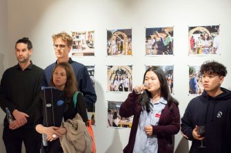 A photo of five people standing in front of a gallery wall, drinks in hand. They appear to be listening as someone beyond the camera speaks. 