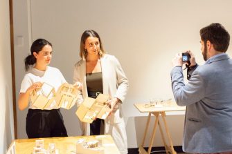 Two woman stand side-by-side, both holding up three-dimensional architectural models and looking towards a man taking a photo of them. 