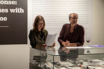 Keith Gallasch and Virginia Baxter speaking behind glass display cabinet containing printed editions of magazine