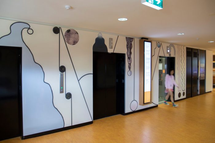 An abstract mural surrounding elevator lifts comprised of curving shapes, pale shades of purple, cream, blue, and textured wall decals