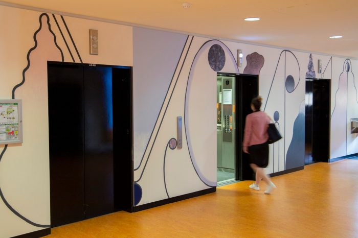 An abstract mural surrounding elevator lifts comprised of curving shapes, pale shades of purple, cream, blue, and textured wall decals