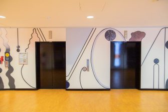 An abstract mural surrounding elevator lifts comprised of curving shapes, pale shades of purple, cream, blue, and textured wall decals