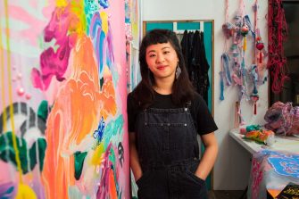A young woman with dark hair and black clothing stands next to a multicoloured abstract painting. A busy workspace is pictured in the background
