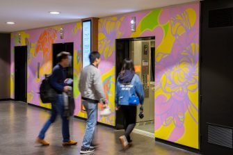 Three students walking toward a multicoloured abstract mural surrounding elevator lifts