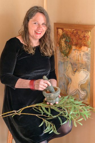 A woman in a black dress with a leafy branch in her lap sits alongside a painting while holding a mortar and pestle.