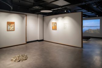 A dimly lit room with tex and small colourful paintings on the walls and a pile of oyster shells in the centre of the grey floor.