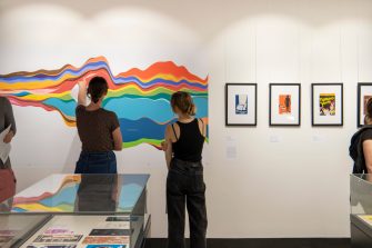 Two women look at a colourful chart with curving rainbow lines. To the right hangs a series of small framed artworks.