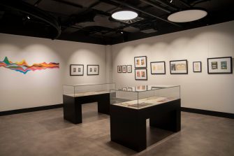 Two glass display cases sit in the centre of a room, a rainbow-coloured line chart on left, artworks in black frames hang on the right.