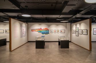 Two glass display cases sit in the centre of a room, a rainbow-coloured line chart on left, artworks in black frames hang on the right.