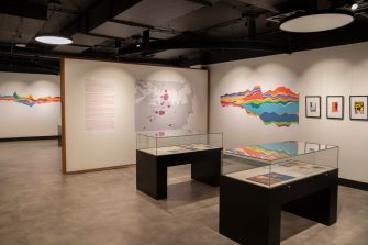 Two glass display cases sit in the centre of a room, a rainbow-coloured line chart on left, artworks in black frames hang on the right.