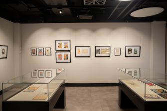 Two glass display cases sit in the centre of a room with artworks in black frames hang in a row on the wall