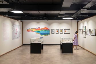 Two glass display cases sit in the centre of a room, a woman in a purple dress looks at artworks in black frames hang on the wall.