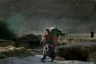Photo of a person in flowing clothing carrying a child in front of them, with a large woven basket on their back.