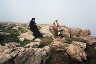 A man and a woman in Iranian clothing sit on rocks a top a hill with a light mist in the background.